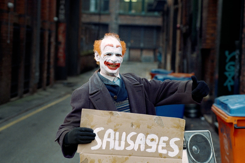 Photograph of a clown holding a sign with the word sausages painted in white